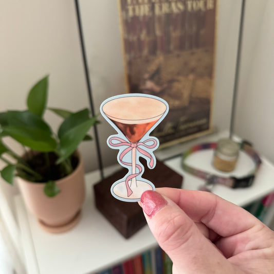 Hand holding a decorative hourglass-shaped sticker with a pink ribbon in front of a shelf with a plant and poster.